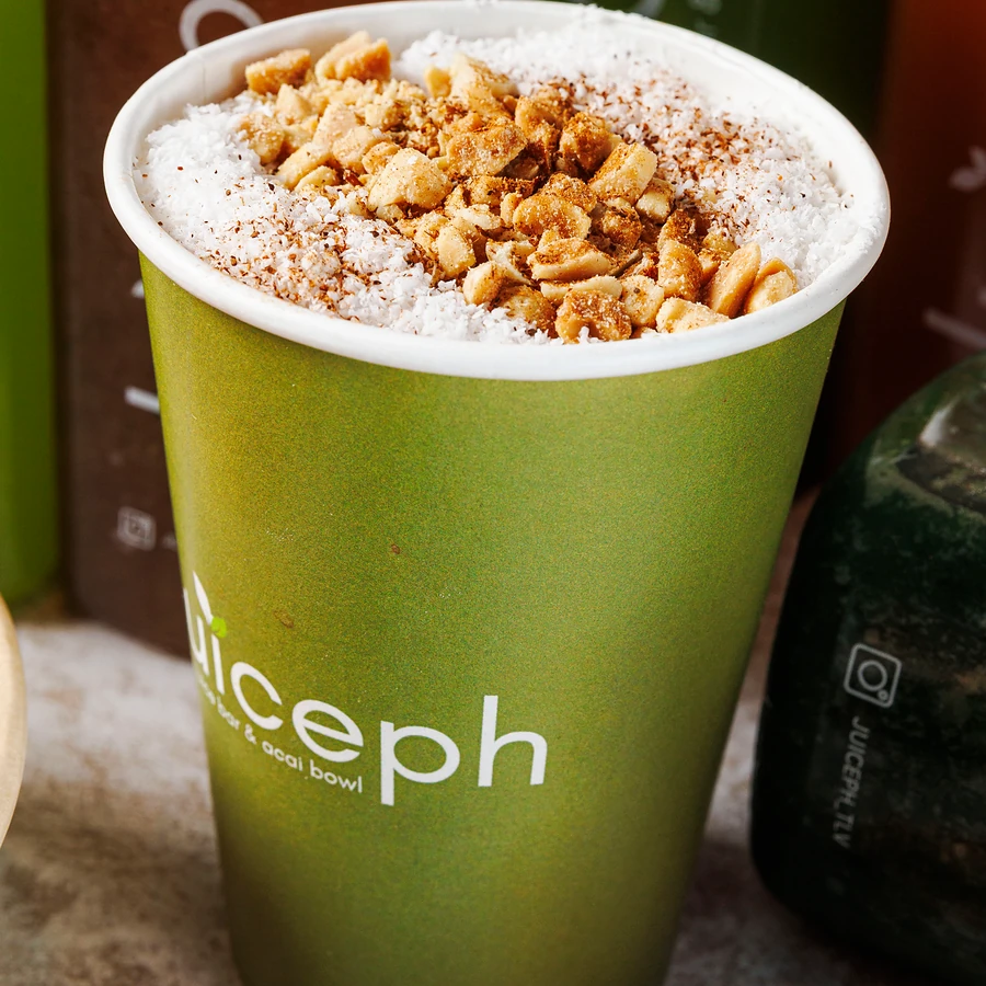 כוס של JUICEPH עם קרם קוקוס, קינמון ובוטנים קצוצים, ומאחור בקבוקי מיץ