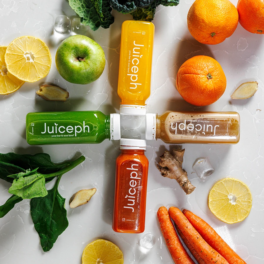 בקבוקי מיץ של JUICEPH בצורת פלוס, מסביב תפוזים, לימונים, תפוח ירוק, גזר, ג׳ינג׳ר ועלים ירוקים